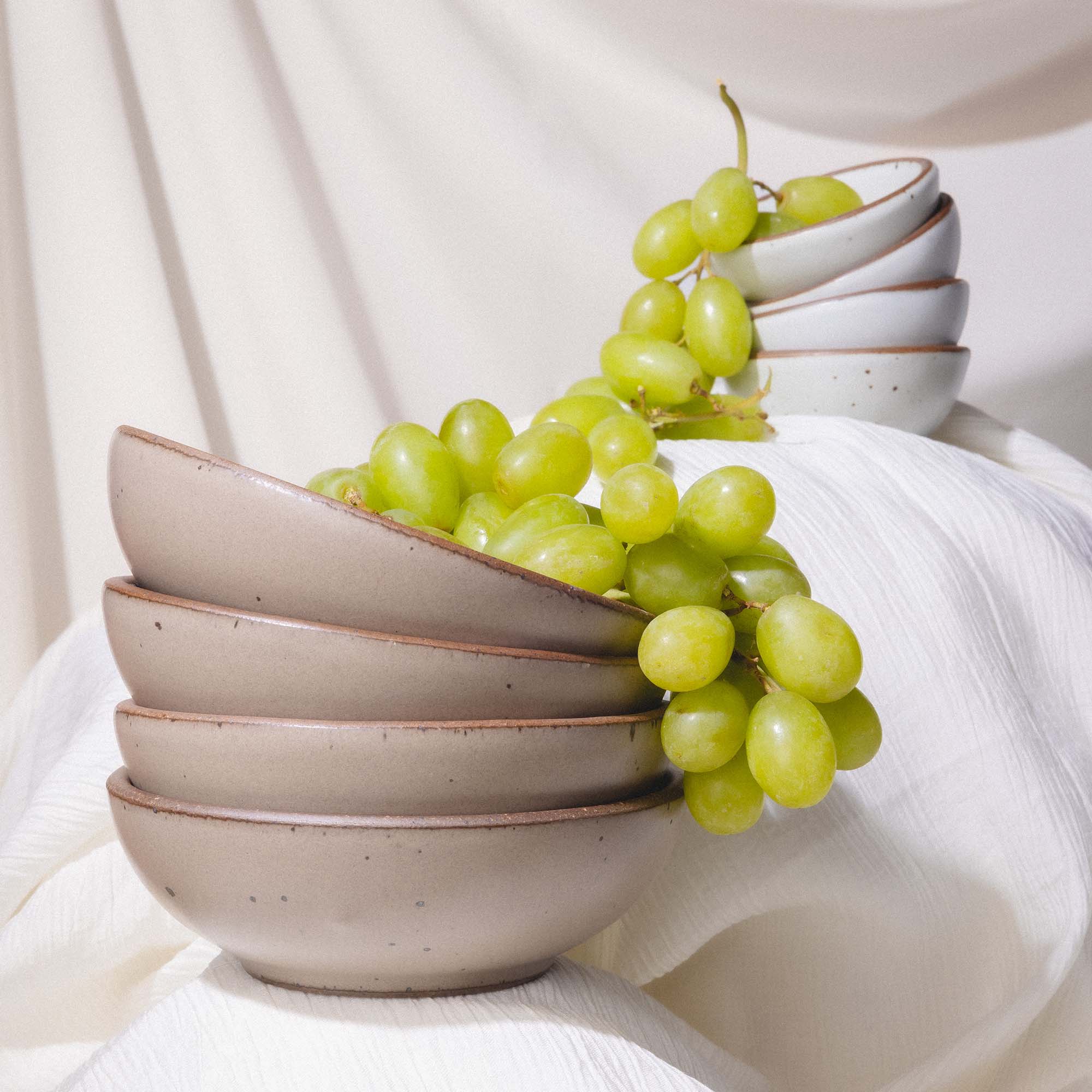A stack of 4 ceramic bowls in a muted tan color with a bundle of green grapes falling out of the top.