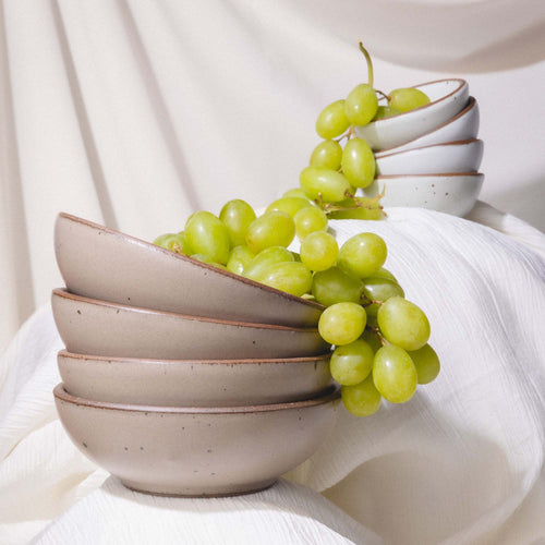A stack of 4 ceramic bowls in a muted tan color with a bundle of green grapes falling out of the top.