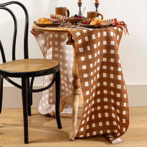 Brown crosshatch table runner draped over a small table set with breakfast pastries, fruit, and mugs, beside a black chair.