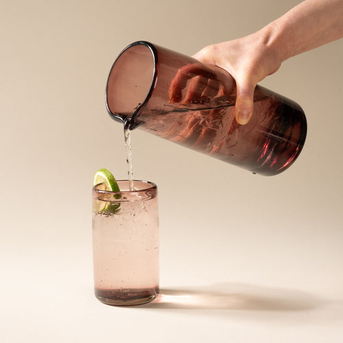 A hand holds a light plum tall transparent glass pitcher with small spout at the top, and pours a drink into a tall tumbler.