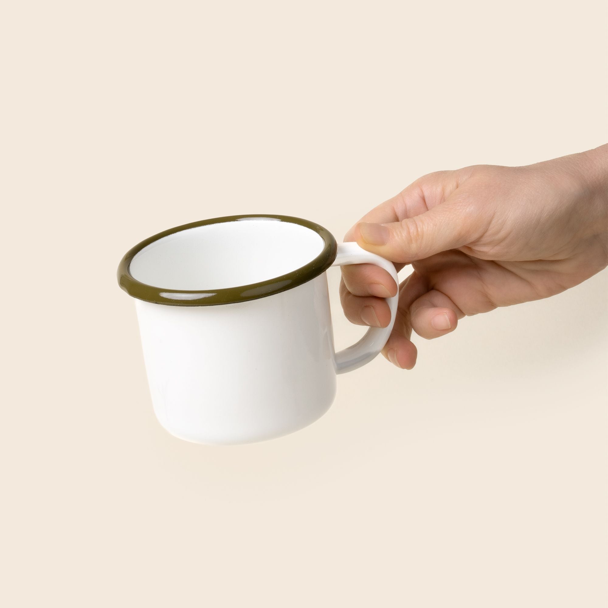 A hand holds out a white enamel mug with an olive green rim and handle, shown against a neutral beige background.