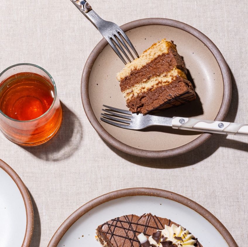 A small slice of cake on a ceramic cake plate in a pale light brown color with sophisticated forks and a glass of red juice.