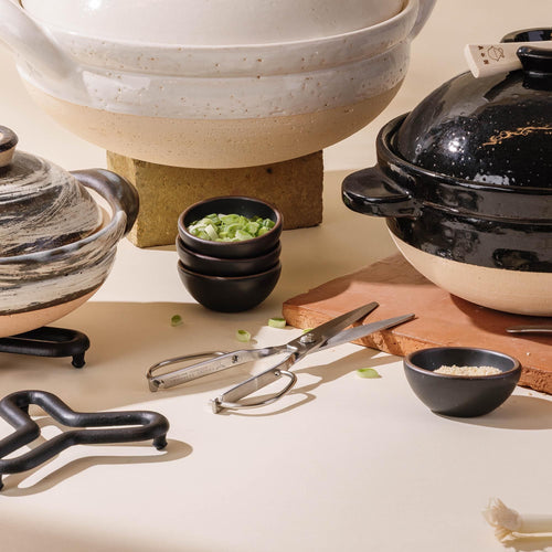 Stacked black mini ceramic bowls with green onions sit beside kitchen scissors, alongside donabes on a neutral surface.