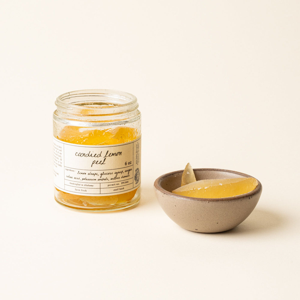 An open jar of candied lemon peel beside a small ceramic bowl holding glossy slices of the preserved fruit.