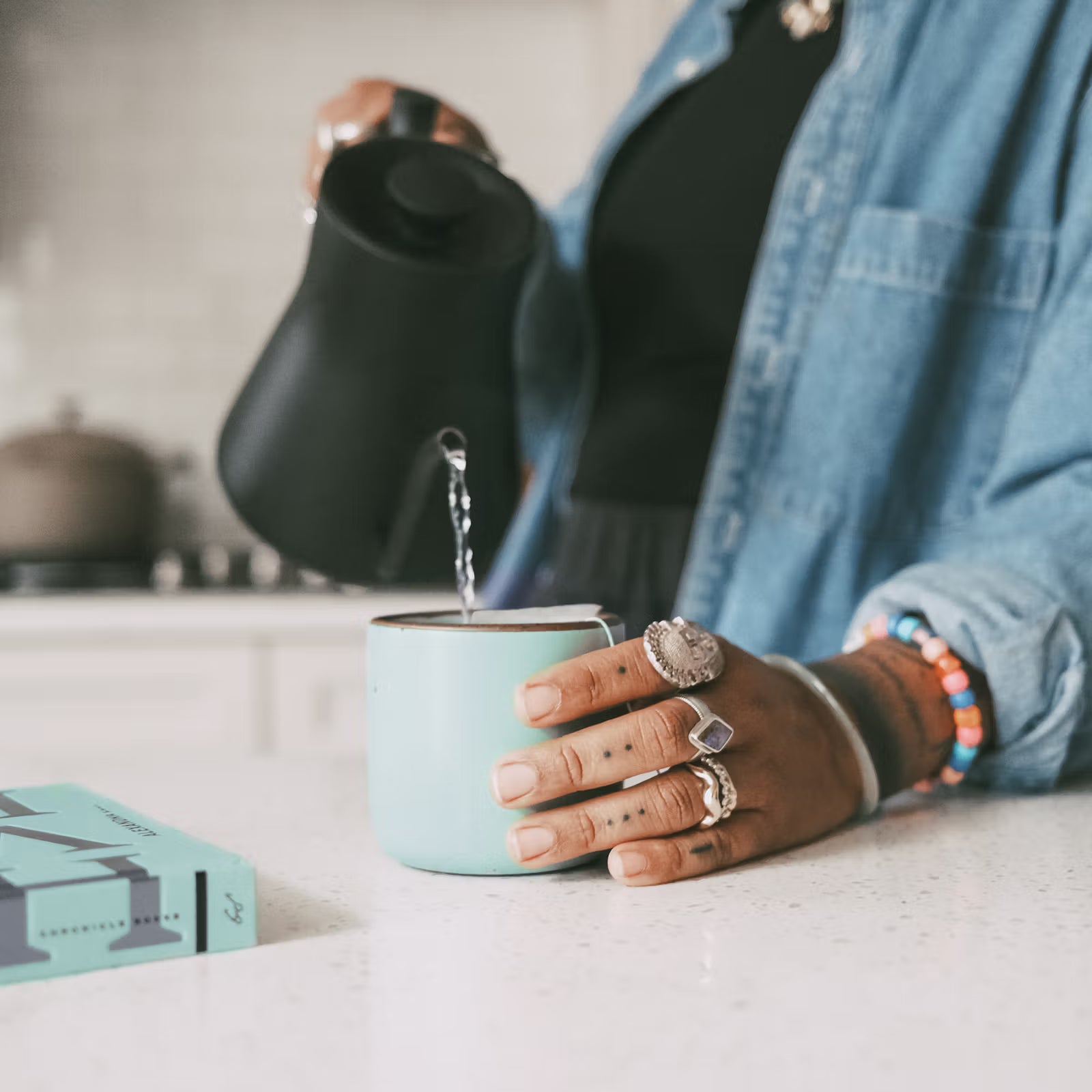 A person is pouring water from a matte black kettle into a light blue mug on a kitchen countertop. Their hand is adorned with rings and a colorful bracelet, and a book lies nearby.