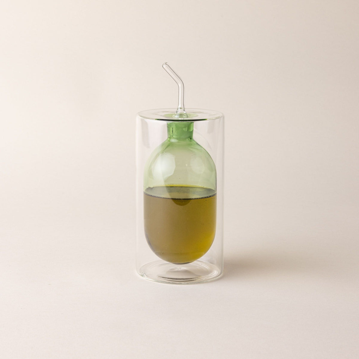 An artful glass cruets in a green color. There is a rounded bulb that acts as a container for the cruet, surrounded by a doubled glass wall. The cruet is filled with oil.