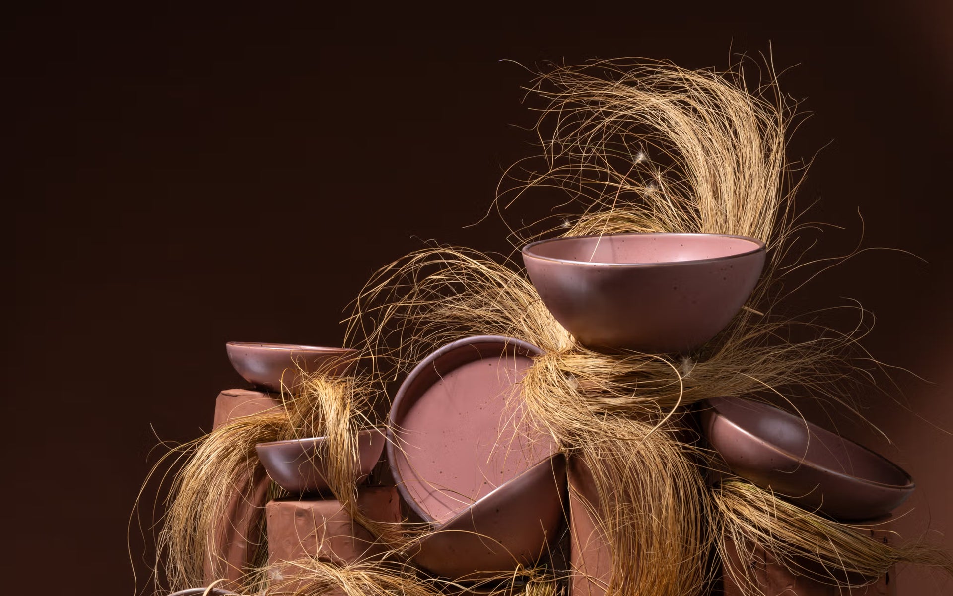 Stack of dusty mauve pink ceramic bowls and bowls piled together with strands of dried grass against a dark background.