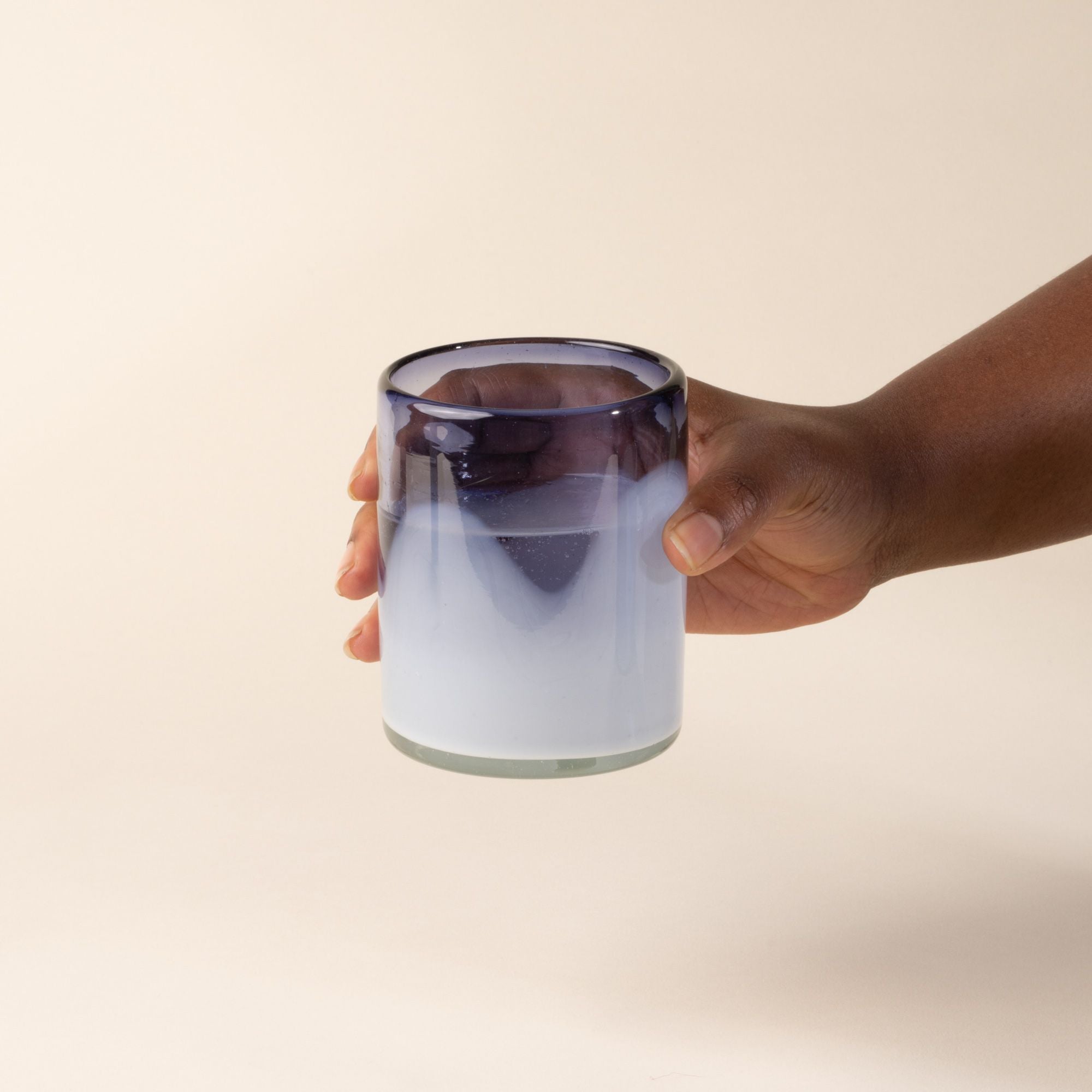 A hand holds out a white and violet handblown glass tumblers with organic wave design, with water.