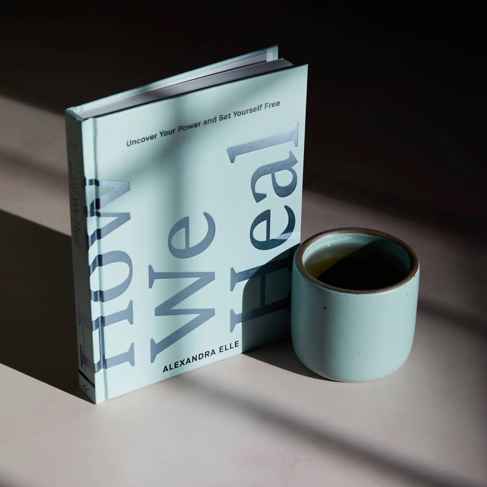 A book titled "How We Heal" by Alexandra Elle stands upright next to a light blue ceramic mug filled with a dark liquid, set on a surface with soft shadows.