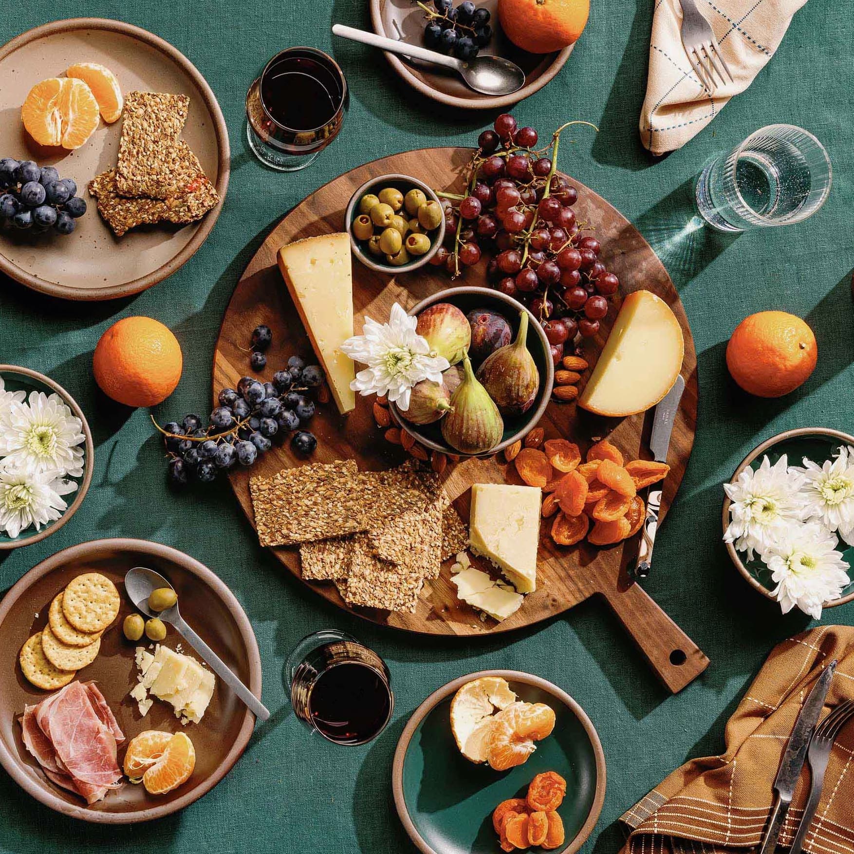 On a table with a teal tablecloth sits a walnut round serving board filled with charcuterie and other foods. Surrounding the board are ceramic plates filled with food, wine glasses, oranges, steel silverware, and napkins.