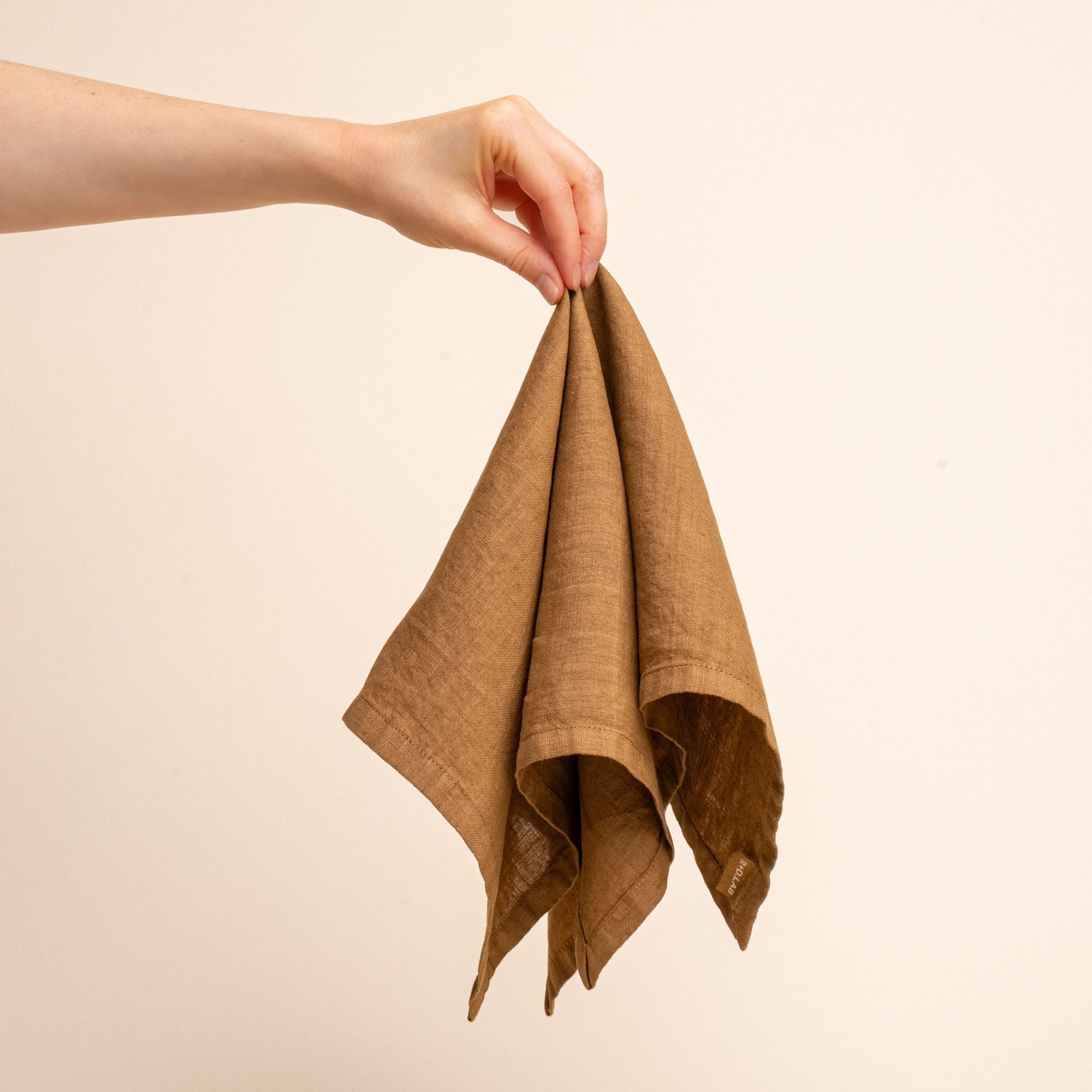A hand holds outs a brown linen napkin against a neutral background.