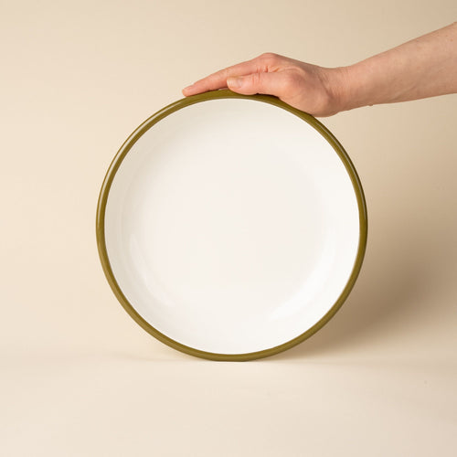 A hand props up an enamel dinner plate in white with an olive green rim.