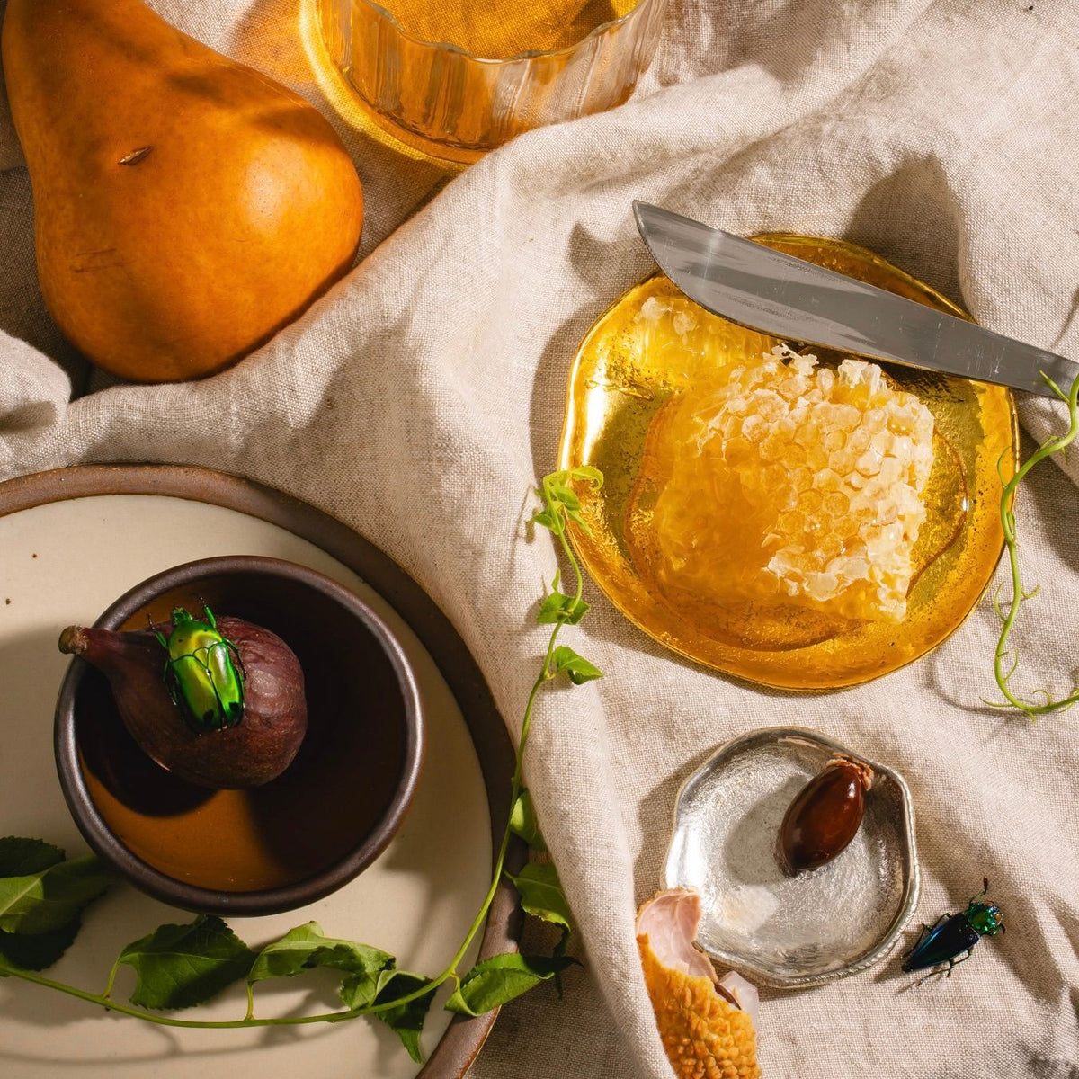 Golden plate with honeycomb and knife, tiny silver dish, fig in a small bowl, pear, and greenery on linen fabric.