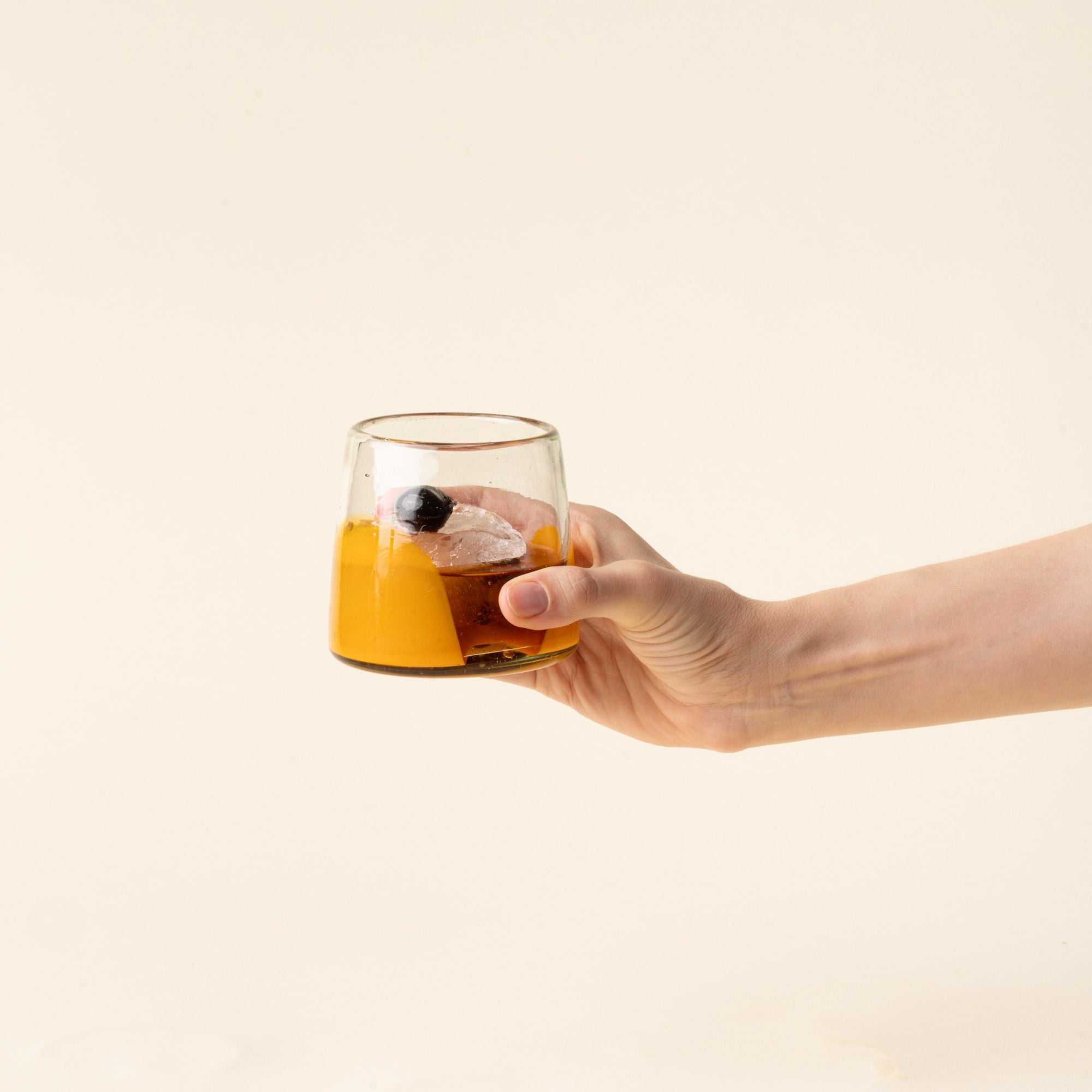 A hand holds out a short clear glasses with orange arch base filled with a cocktail