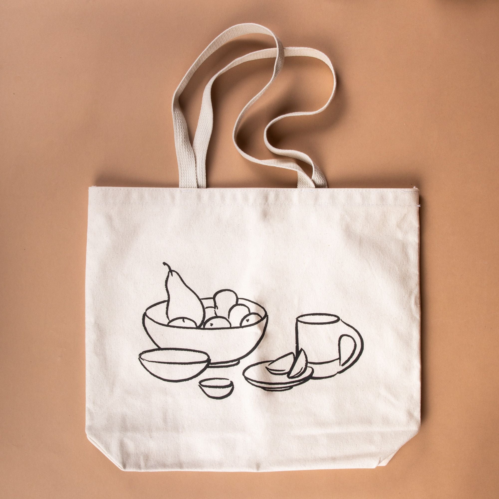 A cream canvas tote has a charcoal illustration on the front of a bowl with fruit, little bowls, plate, and a mug.