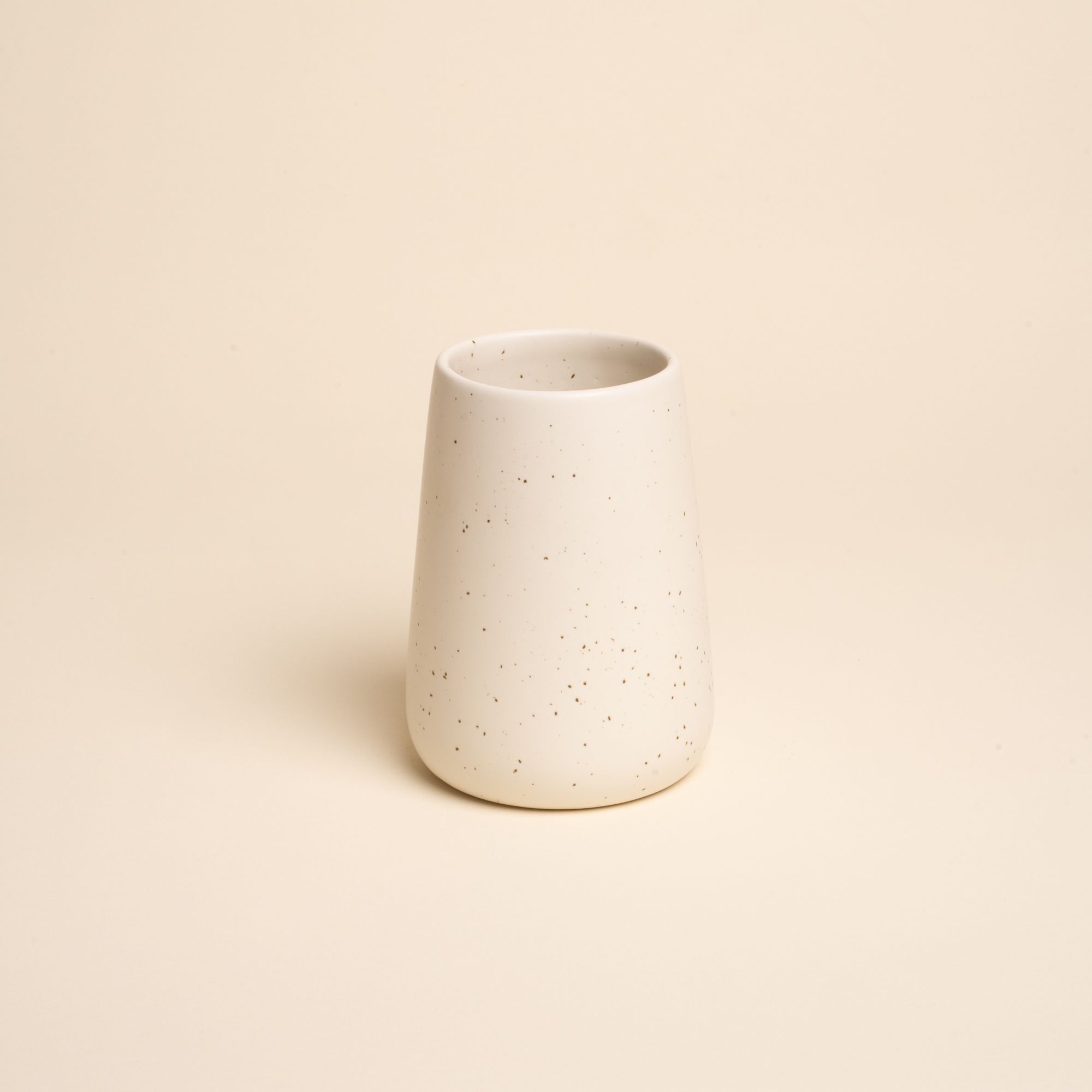 A small minimalist cream-colored tumbler vase with subtle iron speckles stands centered on a soft beige background