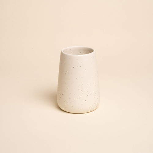 A small minimalist cream-colored tumbler vase with subtle iron speckles stands centered on a soft beige background