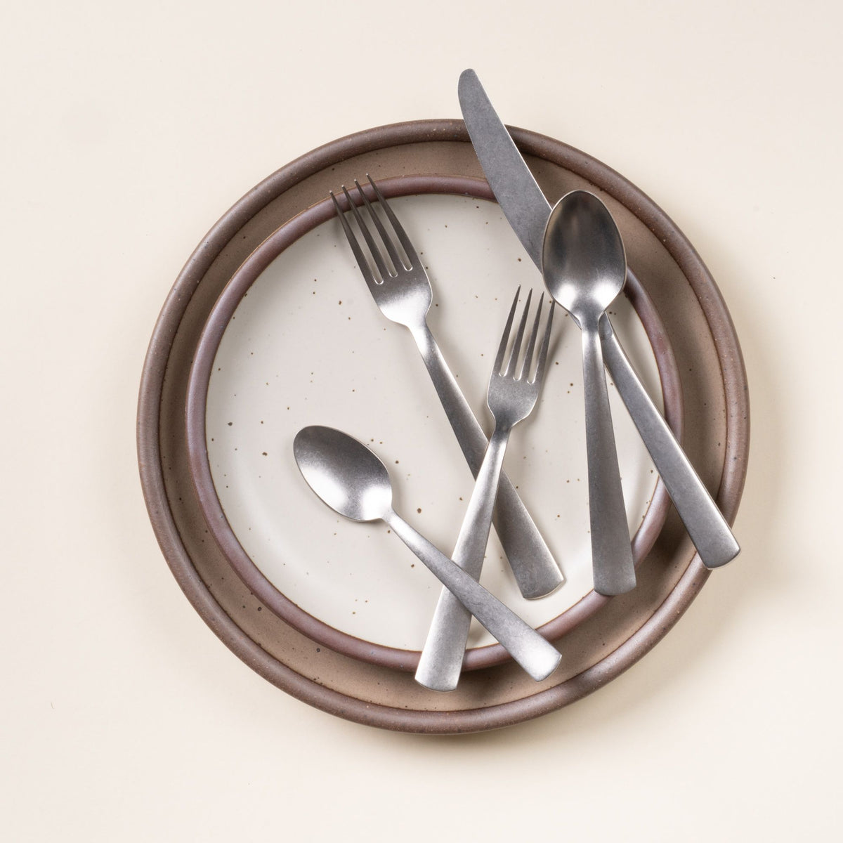 Five matte stainless steel utensils—two forks, knife, tablespoon, and teaspoon—resting on stacked speckled ceramic plates.