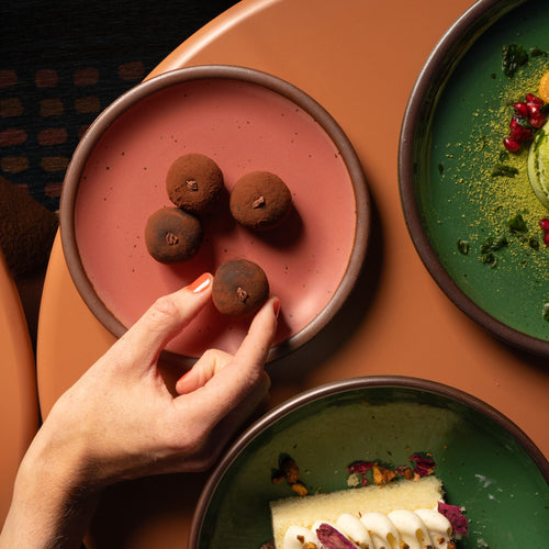 A hand grabs a dessert off a dessert sized ceramic plate in a watermelon pink color featuring iron speckles and an unglazed rim, next to other glossy green plates with desserts.