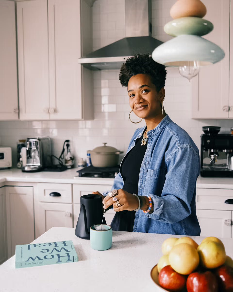 A person stands in a modern kitchen, stirring a drink in a light blue mug beside a black kettle. A book titled "How We Heal" and a bowl of apples are on the counter.