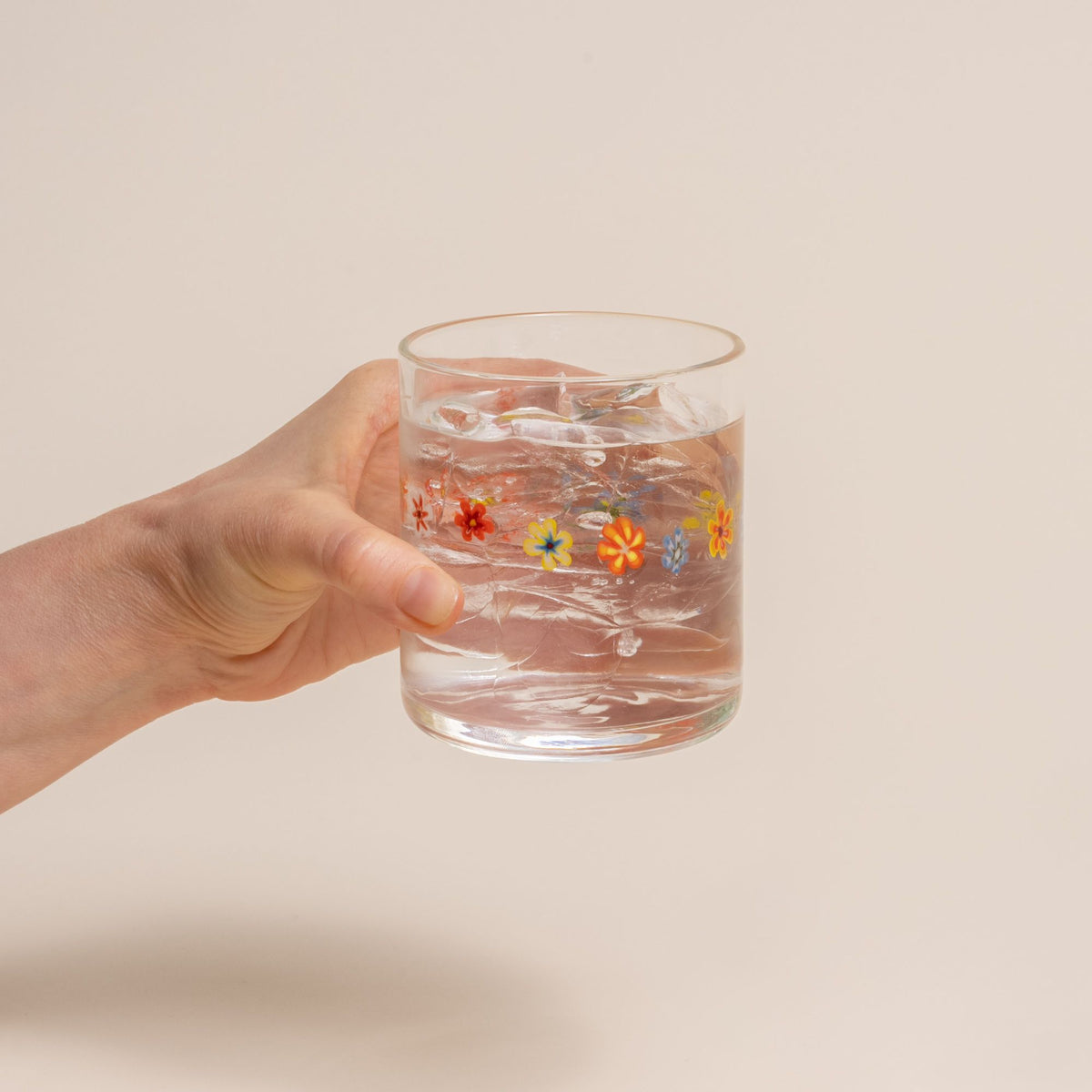 A hand holding a clear glass with a ring of multicolored little flowers, filled with water, on a cream background.