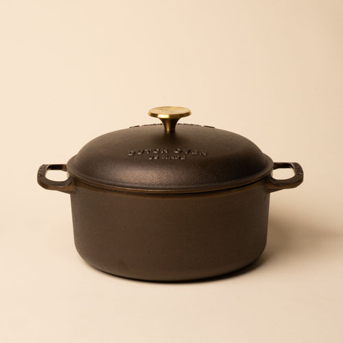 Matte black cast iron Dutch oven with brass knob lid and side handles on a neutral background.