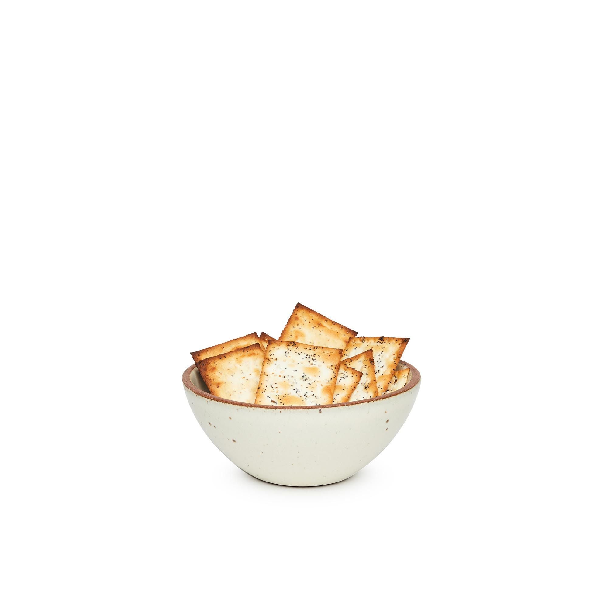 A small dessert sized rounded ceramic bowl in a warm, tan-toned, off-white color featuring iron speckles and an unglazed rim, filled with crackers