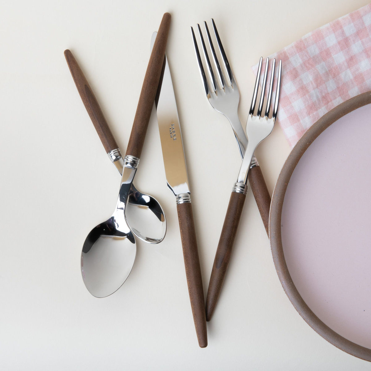 A flatware set of a salad fork, dinner fork, spoon, teaspoon, and knife - all with steel top and simple wood handles. Sitting next to a baby pink plate and a pink and white gingham napkin.