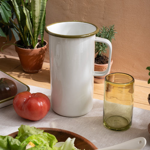 A large enamel pitcher with handle in a white color with an olive green rim sitting on a table with a olive green glass, tomato, and more.