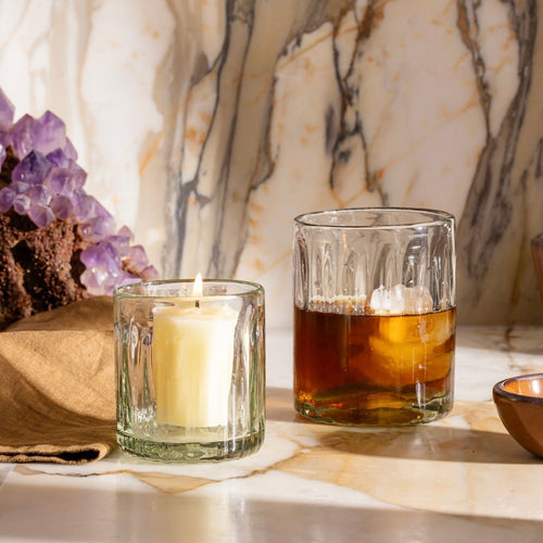 Glass tumbler with a lit candle beside another taller glass filled with iced drink, set on marble with amethyst cluster.