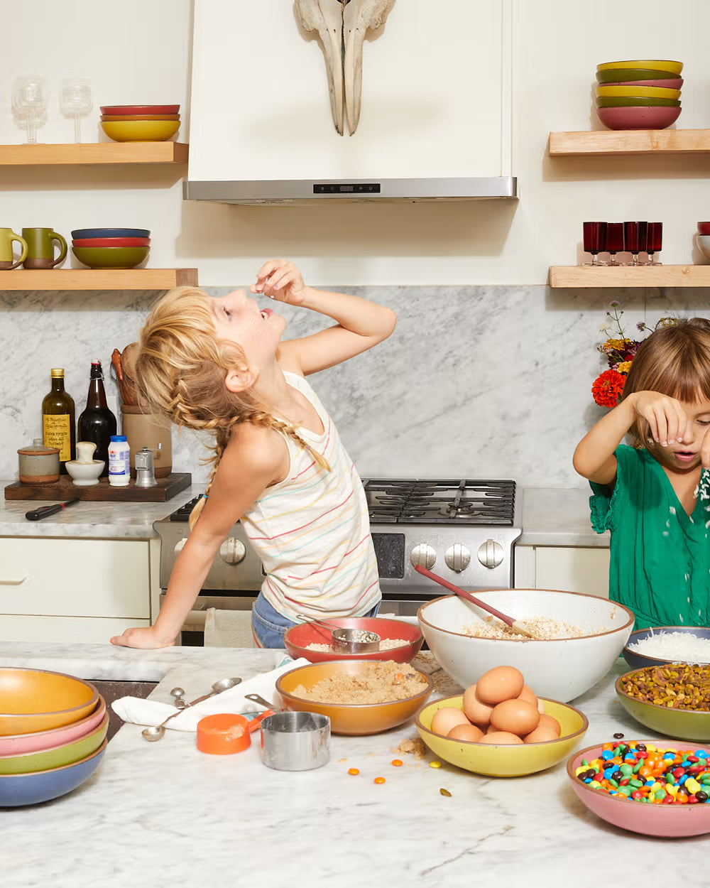 Two children are in a kitchen, engaged in baking activities. Various ingredients, including eggs, candy, and grains, are spread across a marble countertop with stacked colorful bowls and measuring cups nearby.