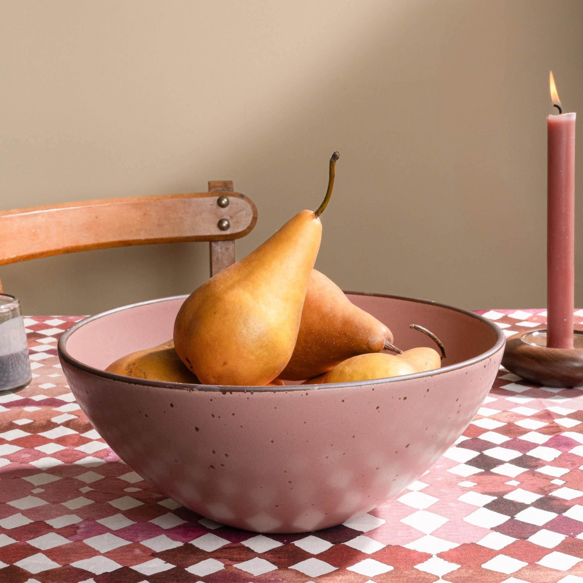 Large ceramic mixing bowl in a dusty mauve pink color filled with pears and citrus on a patterned tablecloth with a wooden chair in the background.
