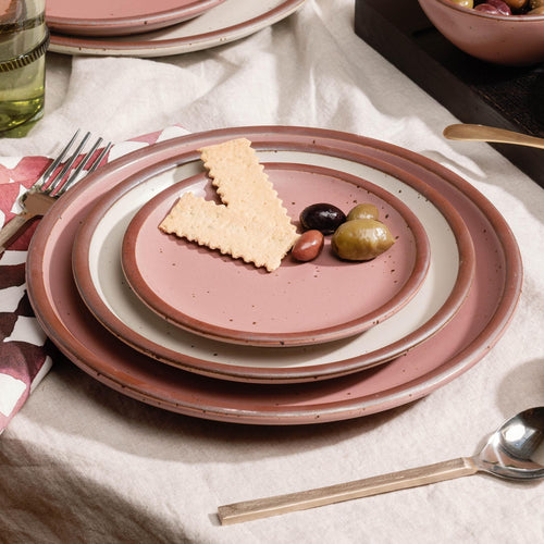 Place setting of stacked dusty mauve pink ceramic plates with crackers and olives on top, set on a white tablecloth with utensils nearby.