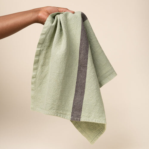 A hand holds the center of a linen towel in a light green color with a dark grey stripe in the middle