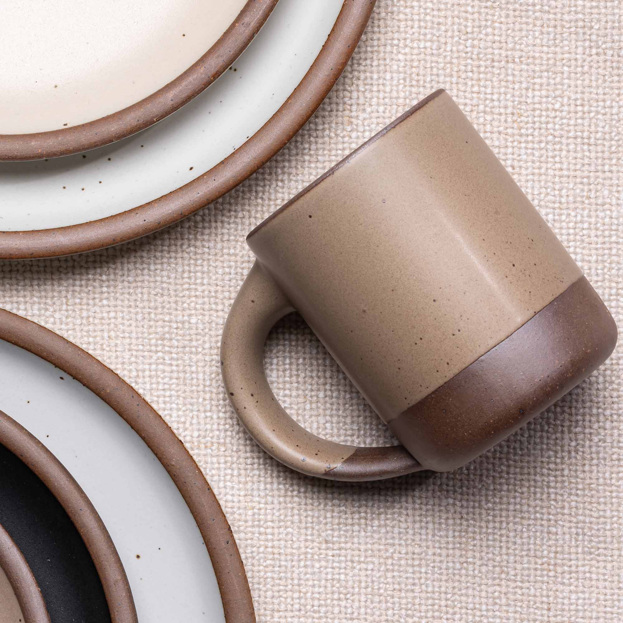A medium sized ceramic mug with handle in a warm pale brown color, surrounded by plates in various sizes.
