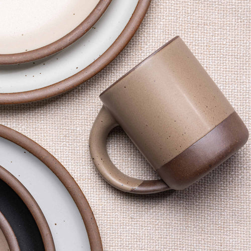 A medium sized ceramic mug with handle in a warm pale brown color, surrounded by plates in various sizes.