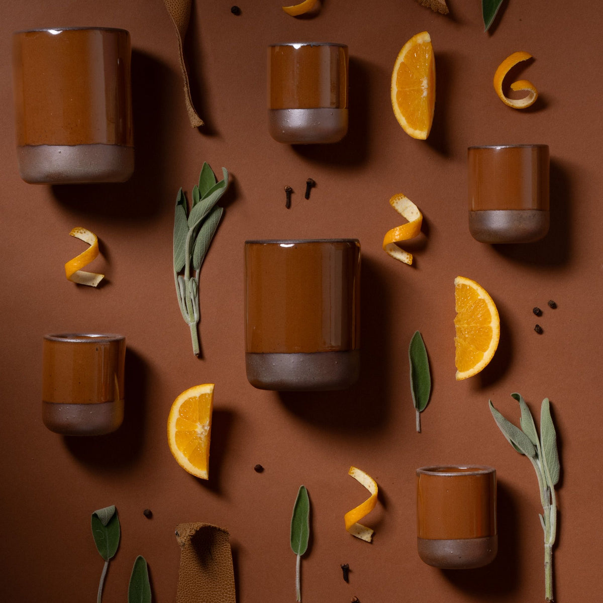 Glossy caramel brown ceramic tumblers arranged with orange slices, peel, sage leaves, and spices on a brown background.