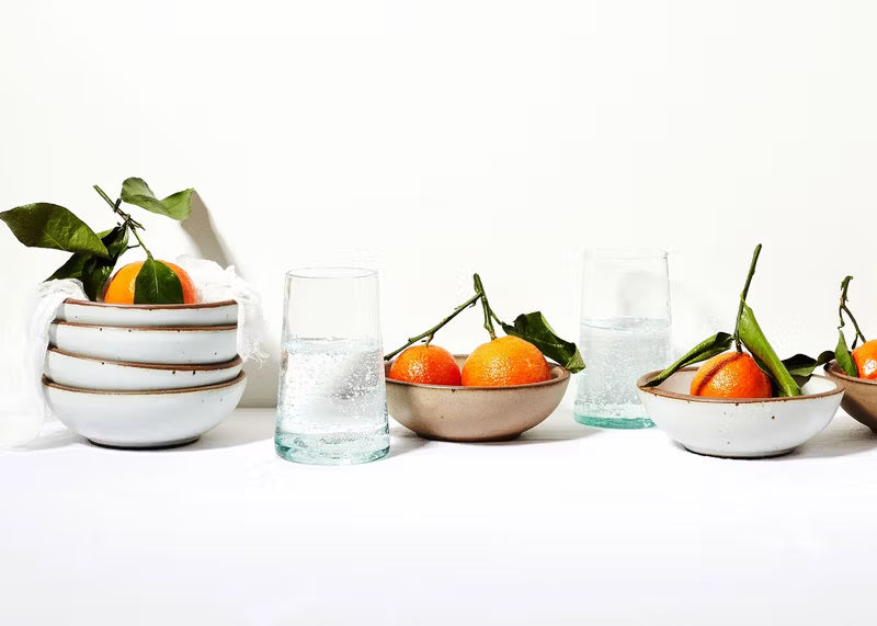Against a white background, are ceramic bowls in white and soft brown colors with oranges, and glasses of water