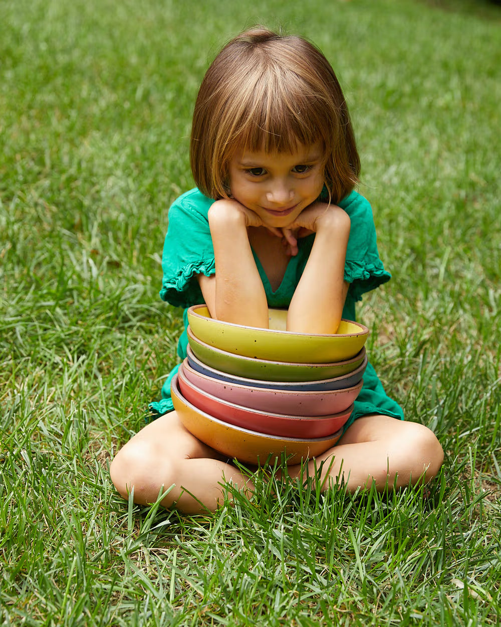 A child in a green shirt sits cross-legged on the grass, holding a stack of colorful ceramic bowls.