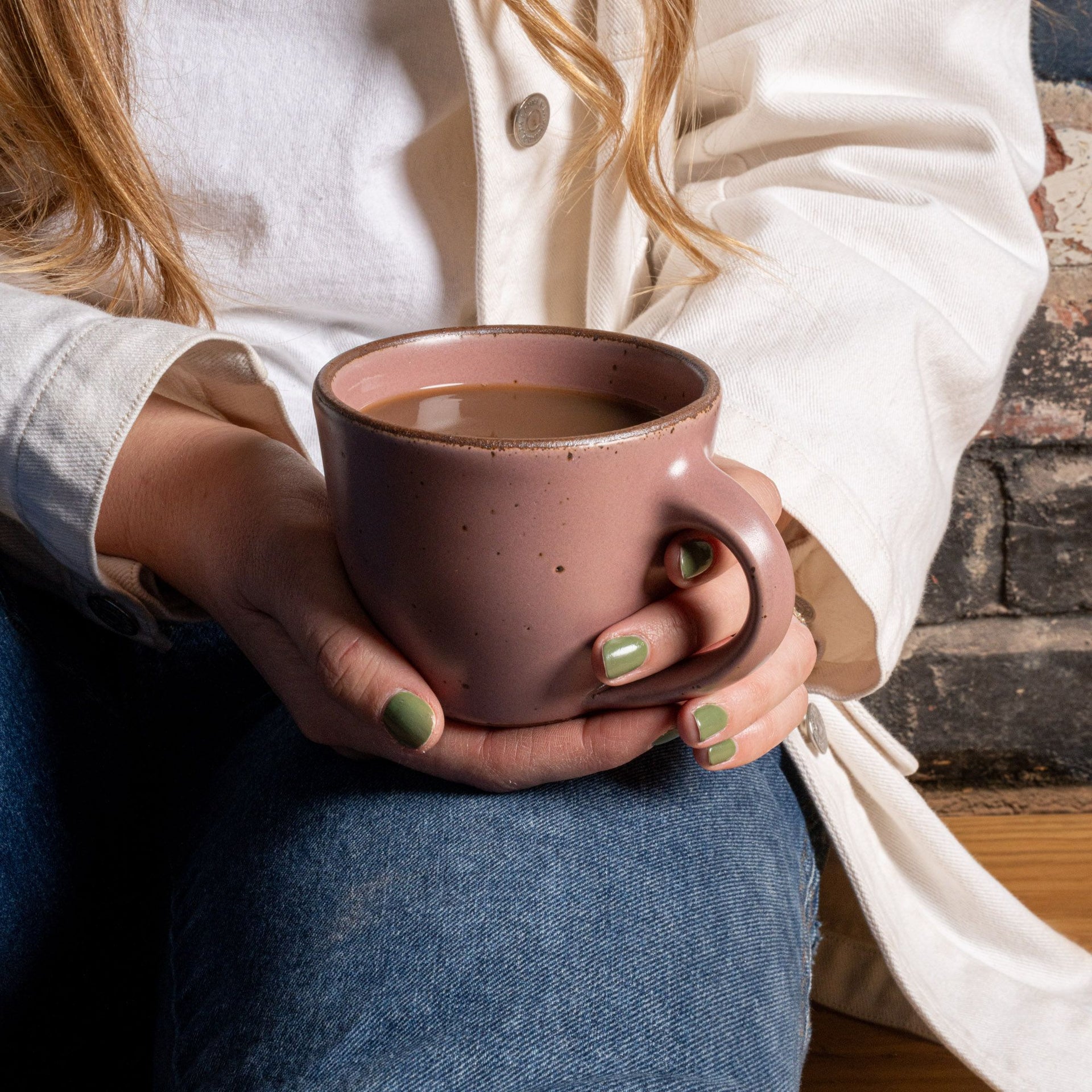 Mallory holds a round stoneware mug with handle, in a dusty mauve pink with iron speckles and an unglazed rim, filled with coffee.