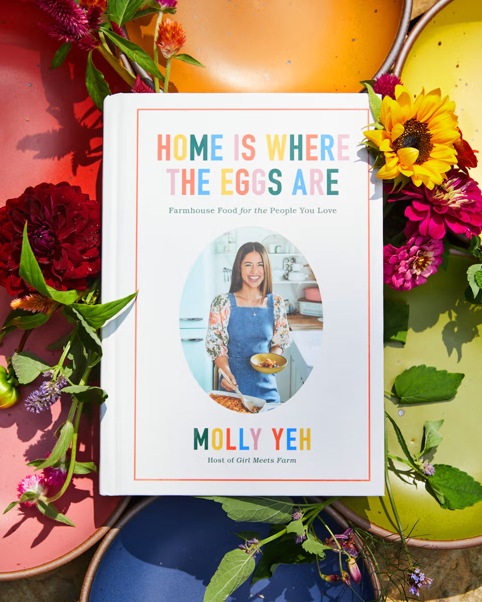 A cookbook titled "Home Is Where the Eggs Are" by Molly Yeh is surrounded by colorful plates and vibrant flowers, creating a cheerful and inviting scene.