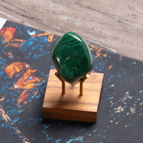 Polished green malachite stone on brass stand with wooden base, displayed against a colorful, abstract book cover.