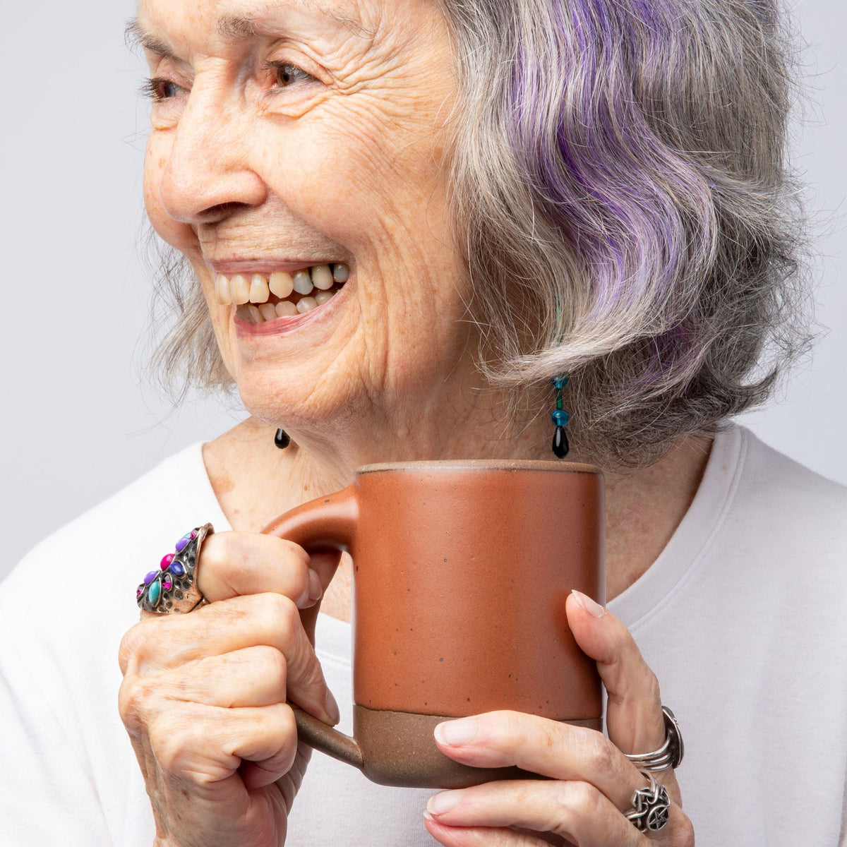 A woman smiling with gray and purple hair holding a medium sized ceramic mug with handle in a cool burnt terracotta color featuring iron speckles and unglazed rim and bottom base.