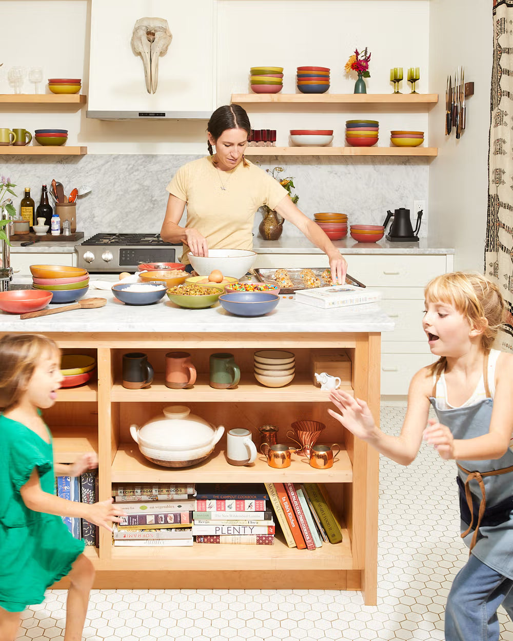 A kitchen scene with a person preparing food surrounded by colorful ceramic bowls on a countertop. Two children play in the foreground, with shelves displaying more ceramics and cookbooks beneath.