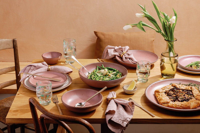 A table filled with ceramic dinnerware in a dusty mauve pink color, a vase with flowers, and dappled pink and green glassware.