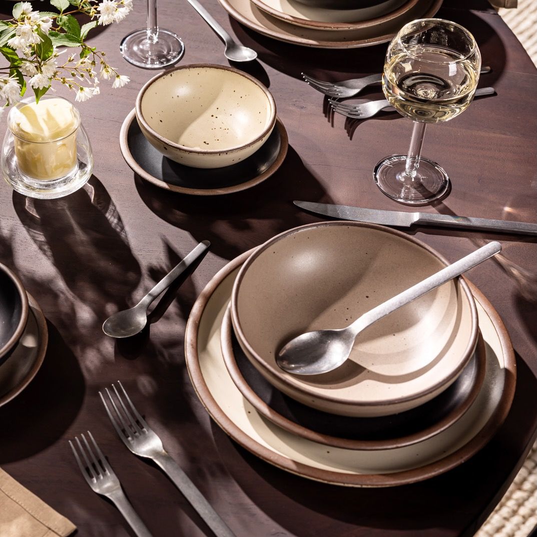 A table filled with ceramic dinnerware in cream, black, and warm pale brown colors, with silverware and wine glasses.