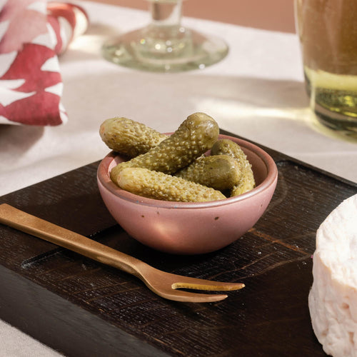 Small ceramic bowl in a dusty mauve pink color of pickles on a wooden serving board with a brass fork and wine glasses in the background.