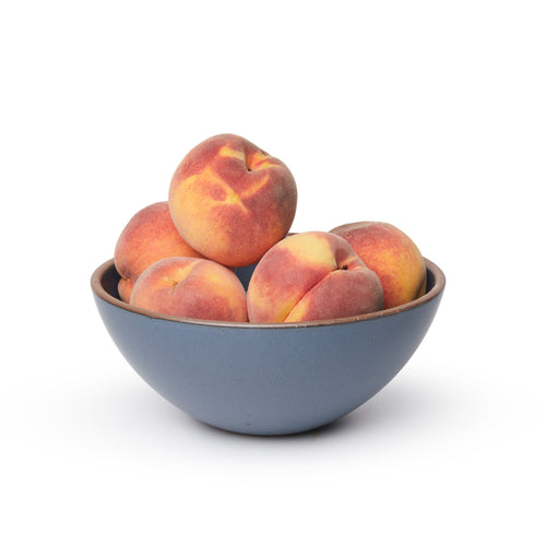 A large rounded ceramic bowl in a cool, medium blue color featuring iron speckles and an unglazed rim, filled with peaches