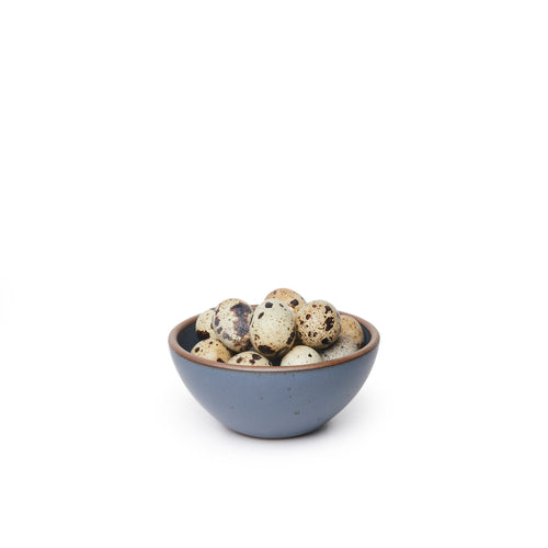 A small dessert sized rounded ceramic bowl in a cool medium blue color featuring iron speckles and an unglazed rim, filled with quail eggs