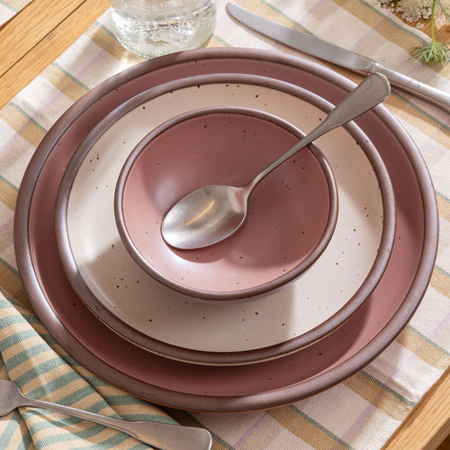Stacked dusty pink and cream ceramic dishware with a spoon on top, set on a plaid tablecloth with a knife and glass nearby.